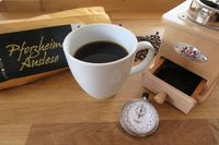 Eine Tasse Kaffee neben einer Kaffeemühle, einer Stoppuhr und einer Tüte Kaffeebohnen.